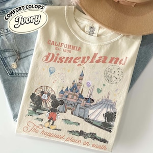 May include: Ivory-colored t-shirt with a Disneyland graphic. The design includes Mickey Mouse, a castle, and the text "Disneyland" and "The happiest place on earth". The shirt is paired with blue jeans and a hat.
