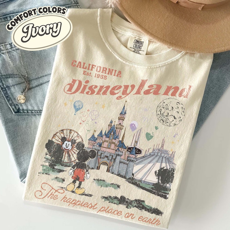 May include: Ivory-colored t-shirt with a Disneyland graphic. The design includes Mickey Mouse, a castle, and the text "Disneyland" and "The happiest place on earth". The shirt is paired with blue jeans and a hat.