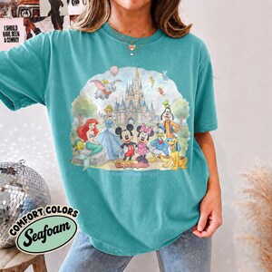 Op de afbeelding: Een blauwgroen t-shirt met ronde hals en een grafische print van Disney-figuren waaronder Mickey Mouse, Minnie Mouse, Donald Duck, Goofy, Pluto, Ari&euml;l en Dumbo voor een kasteel met de tekst Magic Kingdom en Walt Disney World.