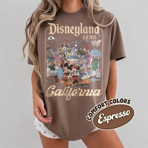 Może przedstawiać: Koszulka w kolorze espresso z grafiką Disneyland California. Projekt zawiera logo Disneyland, rok 1955 oraz postacie z kresk&oacute;wek, takie jak Myszka Miki i Kaczor Donald. Koszulka jest oznaczona jako "Comfort Colors".