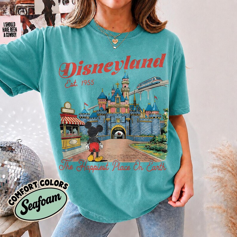 May include: A teal short-sleeve t-shirt featuring a graphic of a castle, a monorail, and Mickey Mouse. The text reads Disneyland Est. 1955 and The Happiest Place On Earth. The shirt has a crew neckline and a relaxed fit with a vintage-style print.