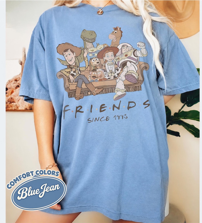 Puede incluir: Camiseta azul claro de manga corta con un gr&aacute;fico de personajes de Toy Story en un sof&aacute; marr&oacute;n. La palabra "FRIENDS" est&aacute; impresa debajo del gr&aacute;fico, con "SINCE 1995" debajo. La camiseta est&aacute; etiquetada como "Blue Jean" en un logotipo de Comfort Colors.