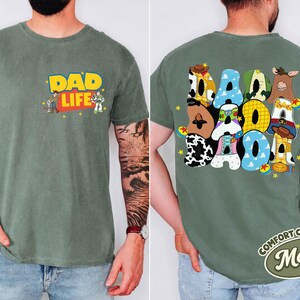 May include: A moss green t-shirt with the words "DAD LIFE" in yellow and red, featuring cartoon characters. The back of the shirt displays the word "DAD" in colorful, patterned letters. A Comfort Colors tag is visible.