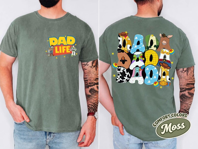 May include: A moss green t-shirt with the words "DAD LIFE" in yellow and red, featuring cartoon characters. The back of the shirt displays the word "DAD" in colorful, patterned letters. A Comfort Colors tag is visible.