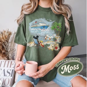 Op de afbeelding: Olijfgroen T-shirt met een strandsc&egrave;ne, inclusief een cruiseschip, palmbomen en Mickey Mouse. Het shirt heeft de woorden "Comfort Colors" en "Moss" in een witte en groene ovaal. Het T-shirt heeft een relaxte pasvorm.
