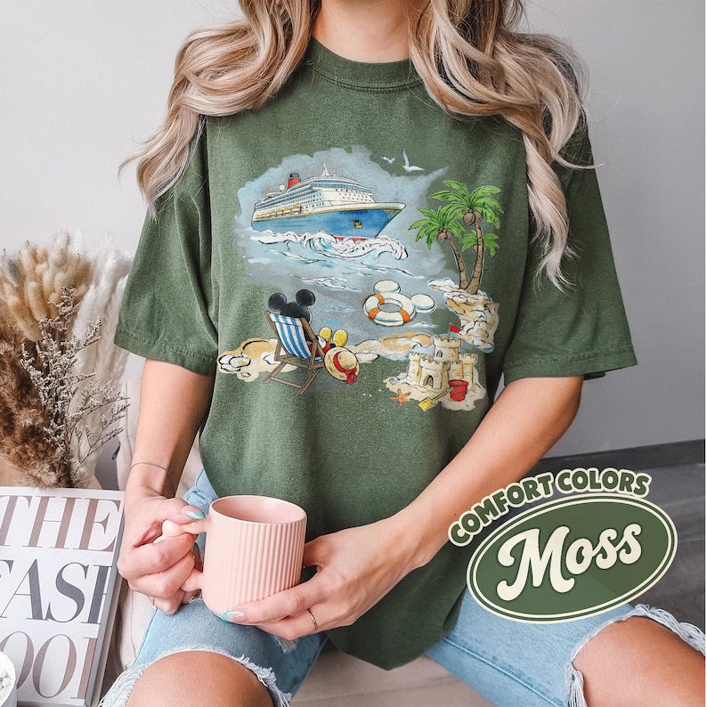 Op de afbeelding: Olijfgroen T-shirt met een strandsc&egrave;ne, inclusief een cruiseschip, palmbomen en Mickey Mouse. Het shirt heeft de woorden "Comfort Colors" en "Moss" in een witte en groene ovaal. Het T-shirt heeft een relaxte pasvorm.