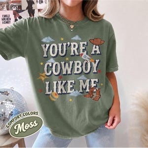 May include: A moss green short-sleeve t-shirt featuring the text "YOU'RE A COWBOY LIKE ME" in a distressed font. The graphic includes illustrations of a brown cowboy hat, a rocket ship, a sheriff star, a ball, and brown cowboy boots.
