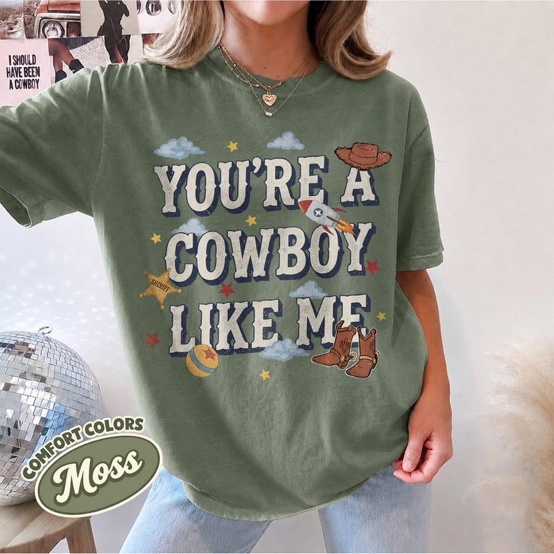 May include: A moss green short-sleeve t-shirt featuring the text "YOU'RE A COWBOY LIKE ME" in a distressed font. The graphic includes illustrations of a brown cowboy hat, a rocket ship, a sheriff star, a ball, and brown cowboy boots.