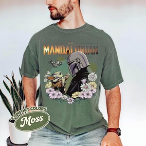 May include: Moss green t-shirt featuring a graphic of The Mandalorian and Grogu surrounded by flowers. The text "THE MANDALORIAN" is printed across the top. The shirt is made of Comfort Colors material.