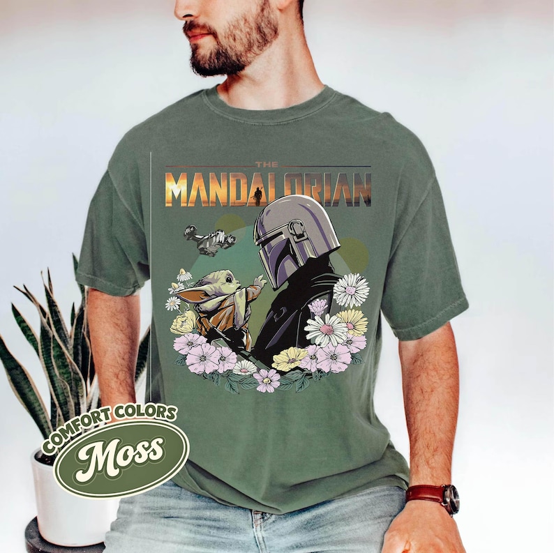 May include: Moss green t-shirt featuring a graphic of The Mandalorian and Grogu surrounded by flowers. The text "THE MANDALORIAN" is printed across the top. The shirt is made of Comfort Colors material.