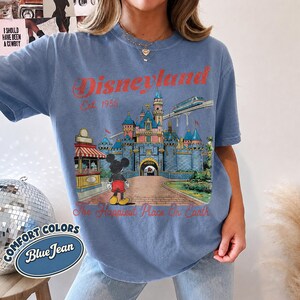 May include: A blue jean colored t-shirt featuring a graphic of the Disneyland castle, Mickey Mouse, and a monorail. The text reads Disneyland Est 1955 and The Happiest Place On Earth in red lettering. The shirt has short sleeves and a crew neckline.