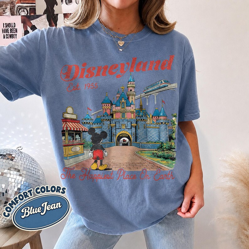 May include: A blue jean colored t-shirt featuring a graphic of the Disneyland castle, Mickey Mouse, and a monorail. The text reads Disneyland Est 1955 and The Happiest Place On Earth in red lettering. The shirt has short sleeves and a crew neckline.