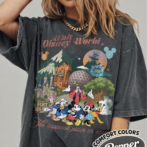 Pu&ograve; includere: T-shirt grigio scuro con una grafica colorata dei monumenti e dei personaggi di Disney World. Il design include il testo "Walt Disney World", "Hollywood Tower" e "The happiest place on earth". La maglietta ha un aspetto vintage.