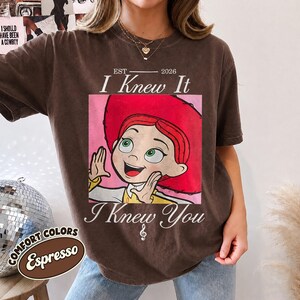 May include: A dark brown short-sleeve t-shirt featuring a graphic of a red-haired character in a red hat. The text reads EST 2026 I Knew It I Knew You with a small musical note symbol. The shirt has a crew neckline and a relaxed fit design.