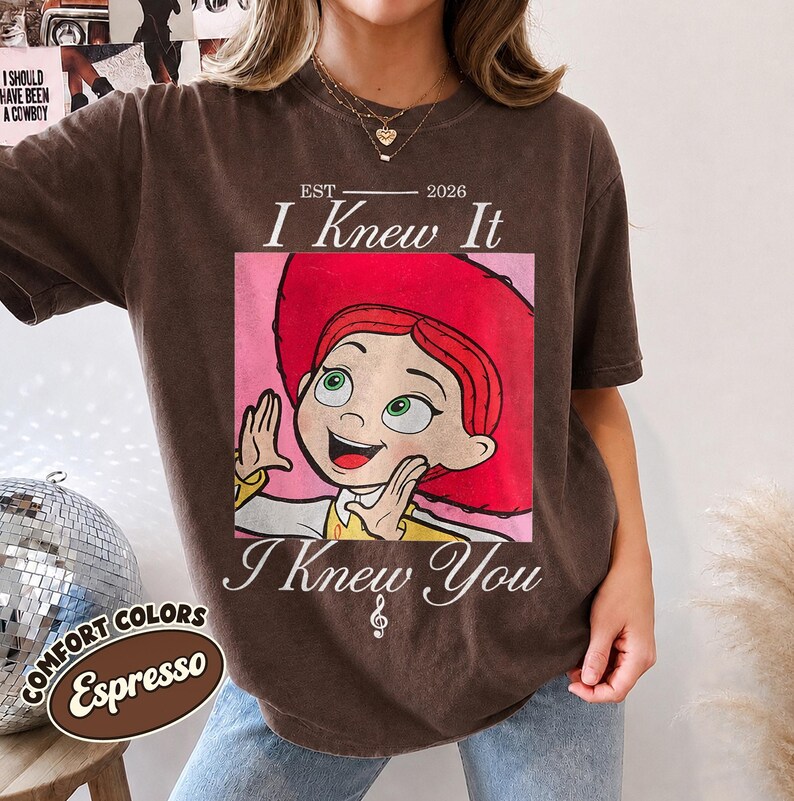 May include: A dark brown short-sleeve t-shirt featuring a graphic of a red-haired character in a red hat. The text reads EST 2026 I Knew It I Knew You with a small musical note symbol. The shirt has a crew neckline and a relaxed fit design.