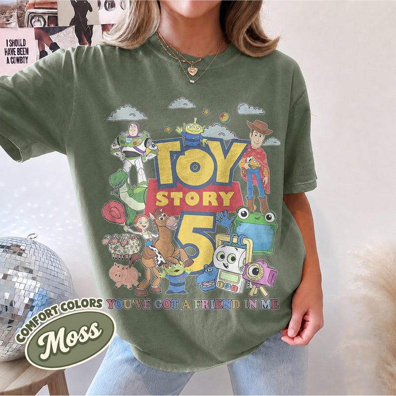 May include: A moss green short-sleeve t-shirt featuring a graphic print of Toy Story characters including Buzz Lightyear, Woody, and Jessie. The design includes the text "TOY STORY 5" and "YOU'VE GOT A FRIEND IN ME" in a distressed, vintage-style font.