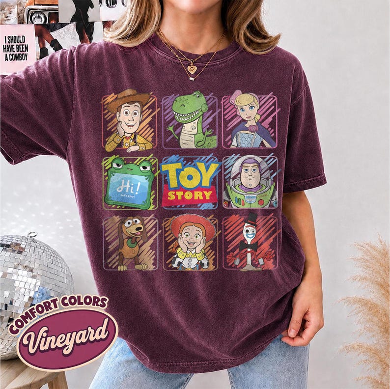 May include: A dark maroon short-sleeve t-shirt featuring a grid of Toy Story characters including Woody, Rex, Bo Peep, a green alien, Buzz Lightyear, Slinky Dog, Jessie, and Forky, with the text Toy Story in the center and Hi! on the alien screen.