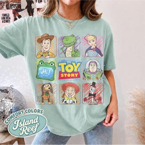 May include: A light green t-shirt featuring a grid of nine squares displaying Toy Story characters including Woody, Rex, Bo Peep, Slinky Dog, Jessie, Forky, and Buzz Lightyear, with a central Toy Story logo and a green tablet displaying the text Hi.