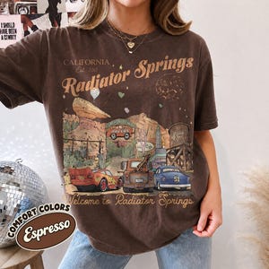 May include: A dark brown short-sleeve t-shirt featuring a graphic print of Radiator Springs with text that reads California Est 2012 Radiator Springs and Welcome to Radiator Springs, depicting a red race car, a brown tow truck, and a blue car.