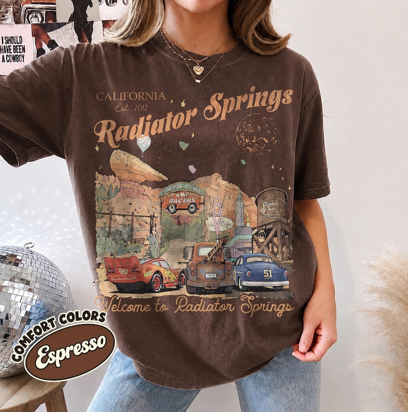 May include: A dark brown short-sleeve t-shirt featuring a graphic print of Radiator Springs with text that reads California Est 2012 Radiator Springs and Welcome to Radiator Springs, depicting a red race car, a brown tow truck, and a blue car.