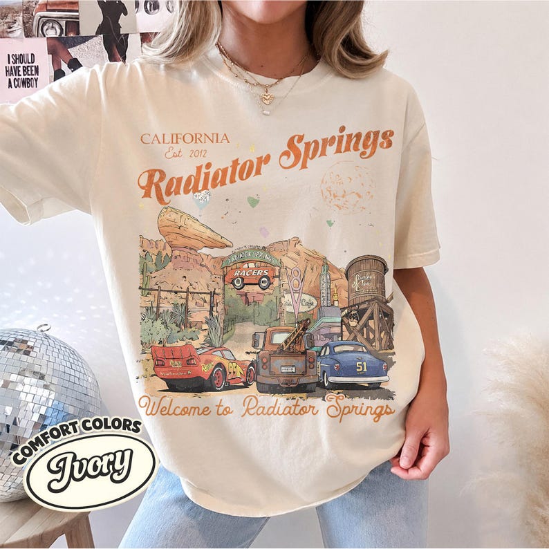 May include: An ivory short-sleeve t-shirt featuring a graphic print of Radiator Springs with text that reads California Est. 2012 Radiator Springs and Welcome to Radiator Springs. The illustration includes desert rock formations and three cars.