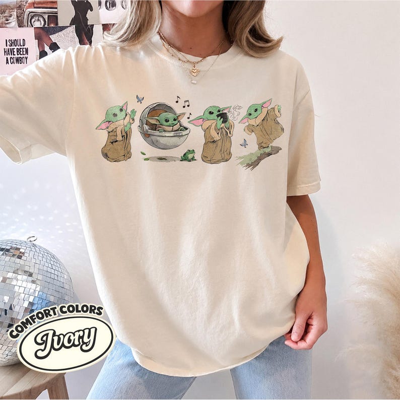 May include: An ivory short-sleeve t-shirt featuring a graphic print of a small green creature in various poses, including sitting in a floating pod, drinking from a cup, and standing, accompanied by small butterflies and musical notes.