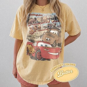 May include: A yellow t-shirt featuring a graphic of Lightning McQueen and Mater from the movie Cars, with the words "Radiator Springs" above them. The shirt has a vintage look and the words "Comfort Colors Butter" are printed on the bottom right.