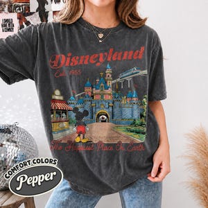 May include: A charcoal gray short-sleeve t-shirt featuring a graphic of a castle, a monorail, and a cartoon mouse. The text reads Disneyland Est. 1955 and The Happiest Place On Earth in red lettering. The shirt has a crew neckline and a relaxed fit.