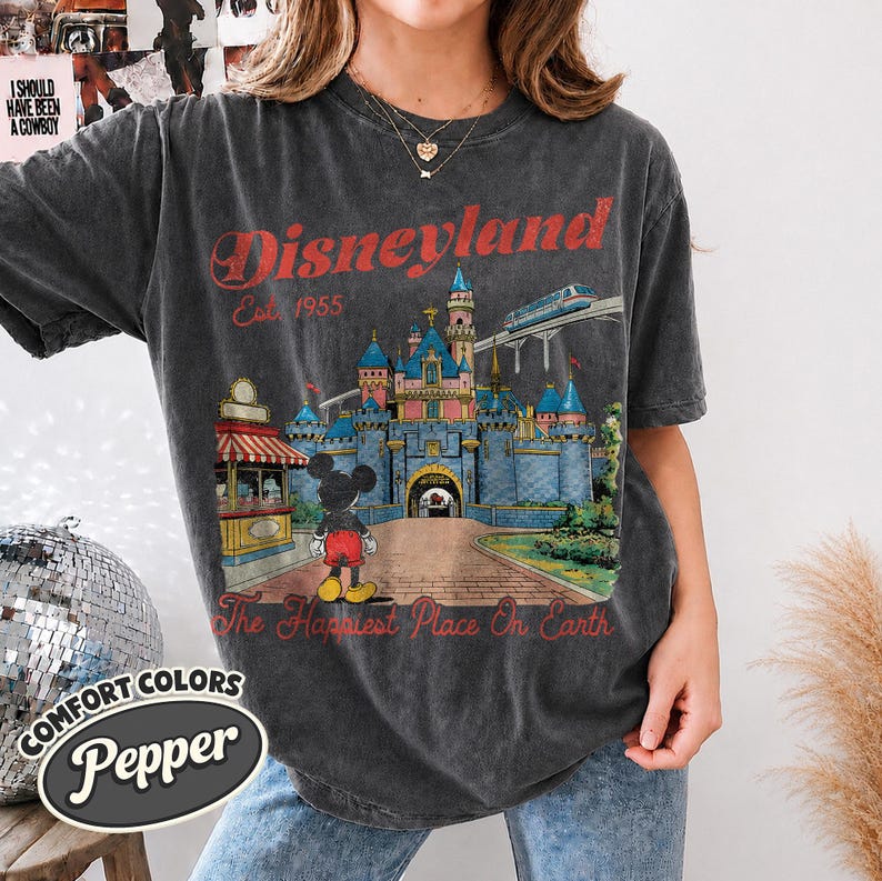 May include: A charcoal gray short-sleeve t-shirt featuring a graphic of a castle, a monorail, and a cartoon mouse. The text reads Disneyland Est. 1955 and The Happiest Place On Earth in red lettering. The shirt has a crew neckline and a relaxed fit.