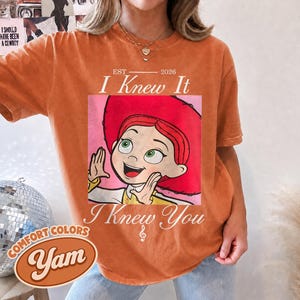 May include: An orange short-sleeve t-shirt featuring a graphic of a cartoon character with red hair and a red hat. The text reads EST 2026 I Knew It I Knew You with a small musical note symbol. The shirt has a crew neckline and a relaxed fit.
