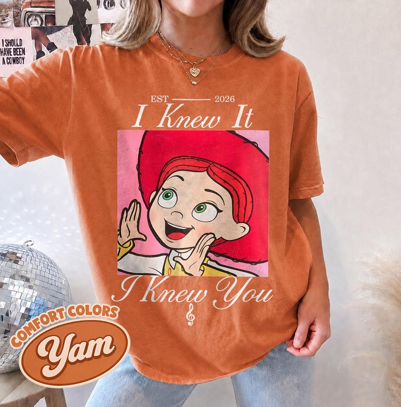 May include: An orange short-sleeve t-shirt featuring a graphic of a cartoon character with red hair and a red hat. The text reads EST 2026 I Knew It I Knew You with a small musical note symbol. The shirt has a crew neckline and a relaxed fit.