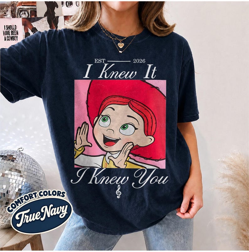 May include: A dark navy blue t-shirt featuring a graphic print of a cartoon character with red hair and a red hat. The text above the graphic reads EST 2026 I Knew It, and the text below reads I Knew You with a small musical note symbol.