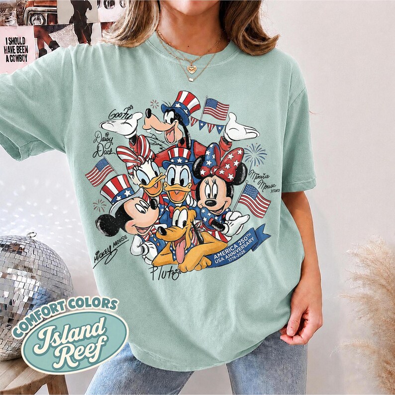 May include: A light green t-shirt featuring a patriotic design with Disney characters Mickey Mouse, Minnie Mouse, Donald Duck, Daisy Duck, Goofy, and Pluto. The design includes American flags and text that reads "America 250th USA Anniversary 1776-2026."