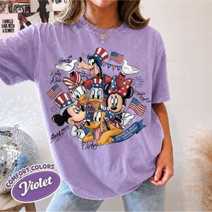 May include: Violet Comfort Colors t-shirt featuring a patriotic design with Disney characters Mickey Mouse, Minnie Mouse, Donald Duck, Daisy Duck, Goofy, and Pluto. The design includes American flags and text celebrating the USA's 250th anniversary.