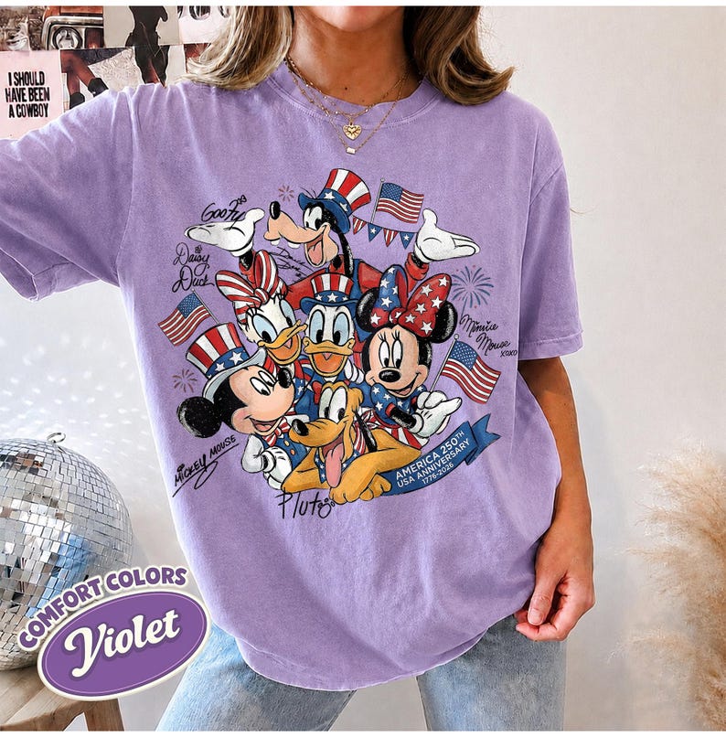 May include: Violet Comfort Colors t-shirt featuring a patriotic design with Disney characters Mickey Mouse, Minnie Mouse, Donald Duck, Daisy Duck, Goofy, and Pluto. The design includes American flags and text celebrating the USA's 250th anniversary.