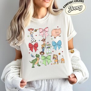 May include: An ivory t-shirt featuring a graphic print of Toy Story characters including Woody, Buzz Lightyear, Jessie, Rex, Hamm, and Slinky Dog, interspersed with pink, red, blue, and green bows and small stars. A Comfort Colors Ivory label is visible.
