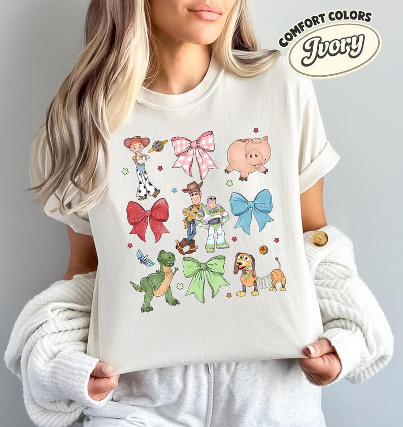 May include: An ivory t-shirt featuring a graphic print of Toy Story characters including Woody, Buzz Lightyear, Jessie, Rex, Hamm, and Slinky Dog, interspersed with pink, red, blue, and green bows and small stars. A Comfort Colors Ivory label is visible.