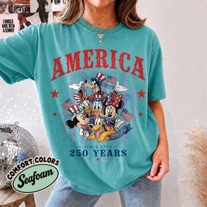 May include: A teal short-sleeve t-shirt featuring a graphic of Disney characters wearing patriotic hats. The text reads AMERICA, SINCE 1776, 250 YEARS, and USA ANNIVERSARY. The design includes red stars and American flags in a distressed style.