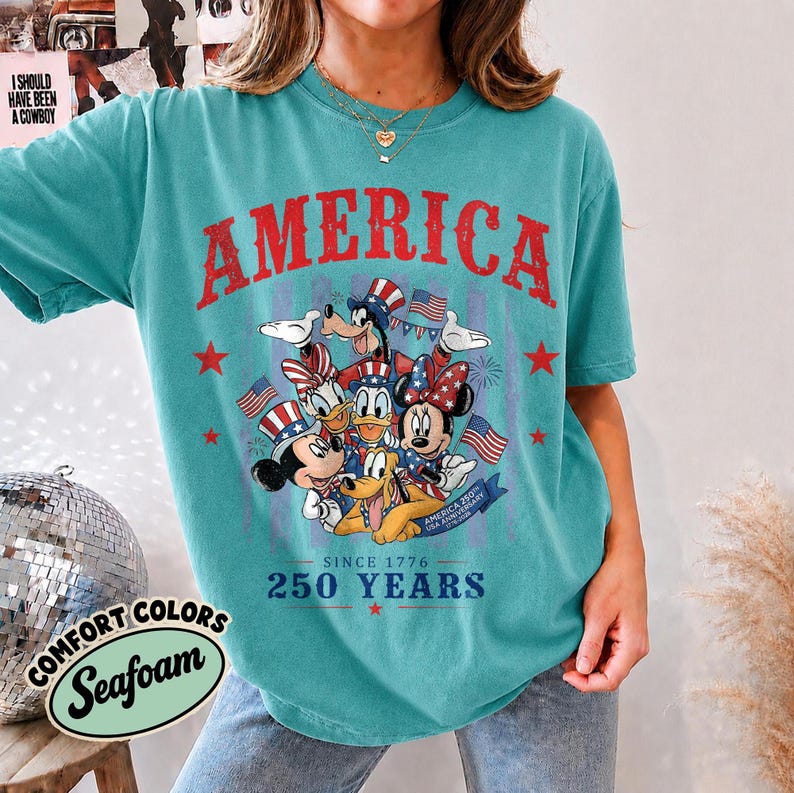 May include: A teal short-sleeve t-shirt featuring a graphic of Disney characters wearing patriotic hats. The text reads AMERICA, SINCE 1776, 250 YEARS, and USA ANNIVERSARY. The design includes red stars and American flags in a distressed style.
