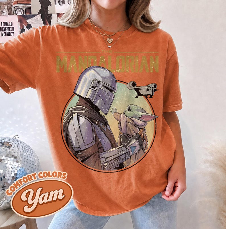 Puede incluir: Una camiseta naranja de manga corta con un estampado gr&aacute;fico de The Mandalorian y Grogu dentro de un marco circular. El texto "THE MANDALORIAN" est&aacute; impreso en una fuente desgastada sobre la ilustraci&oacute;n circular, que incluye una nave espacial.