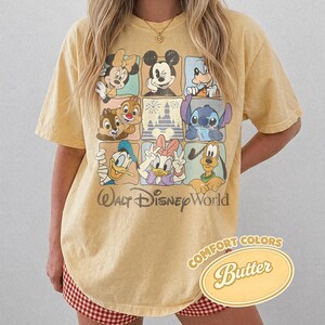 May include: Yellow t-shirt featuring a grid of Disney characters including Mickey Mouse, Minnie Mouse, Donald Duck, and Stitch. The shirt has the text "Walt Disney World" and "Comfort Colors Butter" printed on it.