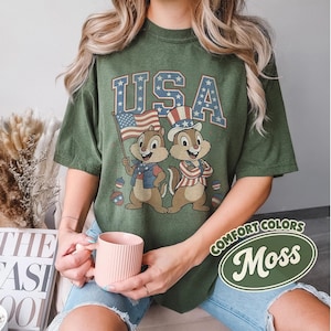 May include: Moss green t-shirt featuring a patriotic design with the word "USA" and cartoon chipmunks holding an American flag and wearing patriotic attire. The shirt is a comfort color.