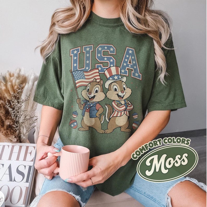May include: Moss green t-shirt featuring a patriotic design with the word "USA" and cartoon chipmunks holding an American flag and wearing patriotic attire. The shirt is a comfort color.
