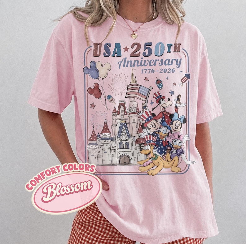 Puede incluir: Camiseta rosa claro con un dise&ntilde;o que celebra el 250 aniversario de EE. UU. El dise&ntilde;o incluye un castillo, fuegos artificiales y personajes de dibujos animados. El texto "USA 250th Anniversary 1776-2026" se muestra en un marco azul. La camiseta tambi&eacute;n tiene el texto "Comfort Colors Blossom".