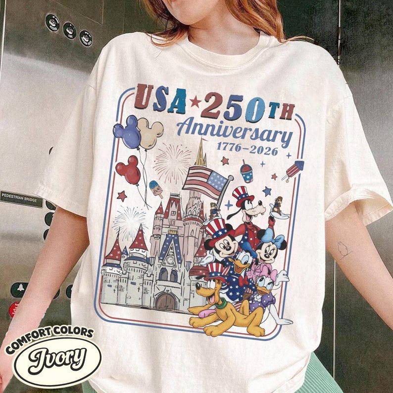 Puede incluir: Camiseta marfil Comfort Colors con un dise&ntilde;o patri&oacute;tico para el 250 aniversario de EE. UU. El gr&aacute;fico incluye a Mickey Mouse, Minnie Mouse, Donald Duck, Daisy Duck, Goofy, Pluto, un castillo y la bandera estadounidense.
