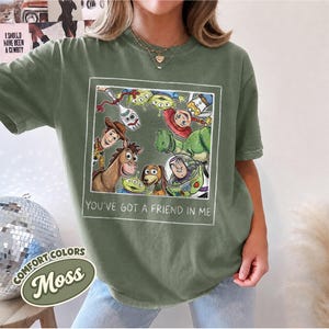 Puede incluir: Camiseta verde musgo con un gr&aacute;fico enmarcado de personajes de Toy Story, incluyendo a Woody y Buzz Lightyear. El texto "YOU'VE GOT A FRIEND IN ME" est&aacute; debajo. La camiseta tambi&eacute;n tiene el texto "COMFORT COLORS" y "Moss".