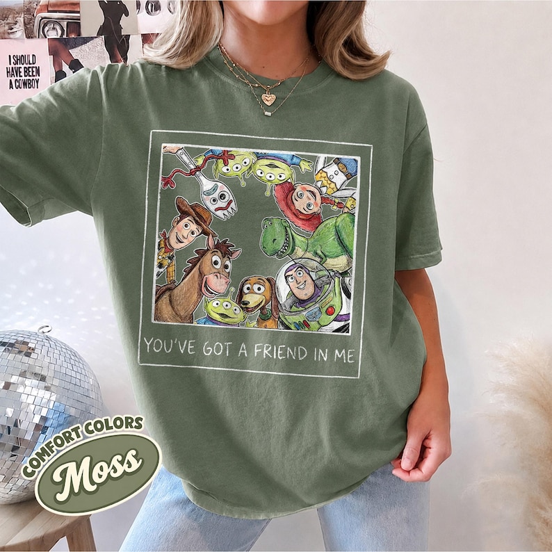 Puede incluir: Camiseta verde musgo con un gr&aacute;fico enmarcado de personajes de Toy Story, incluyendo a Woody y Buzz Lightyear. El texto "YOU'VE GOT A FRIEND IN ME" est&aacute; debajo. La camiseta tambi&eacute;n tiene el texto "COMFORT COLORS" y "Moss".