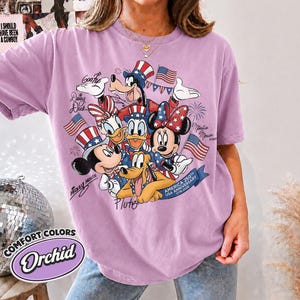 May include: Orchid-colored t-shirt featuring a graphic of Disney characters Mickey Mouse, Minnie Mouse, Donald Duck, Daisy Duck, Goofy, and Pluto in patriotic attire. The design includes American flags and text that reads "AMERICA 250TH USA ANNIVERSARY 1776-2026".