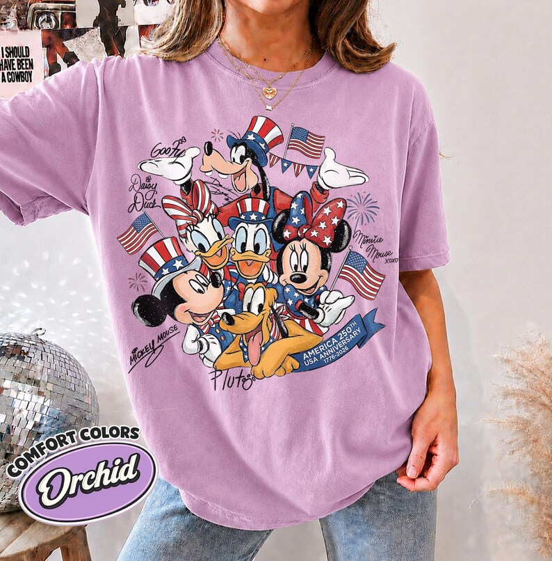 May include: Orchid-colored t-shirt featuring a graphic of Disney characters Mickey Mouse, Minnie Mouse, Donald Duck, Daisy Duck, Goofy, and Pluto in patriotic attire. The design includes American flags and text that reads "AMERICA 250TH USA ANNIVERSARY 1776-2026".