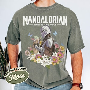 May include: Moss green t-shirt featuring a graphic of The Mandalorian and Grogu surrounded by flowers and butterflies. The text "MANDALORIAN - THIS IS THE WAY" is printed above the graphic. The shirt has a Comfort Colors label.